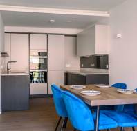 Modern kitchen and dining area with white cabinets, a wooden table set with plates, and bright blue chairs on a wooden floor.