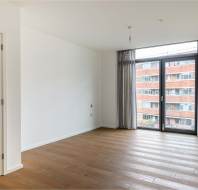 Empty room with wooden floor, white walls, and a large window with sheer curtains. The window offers a view of a brick building outside. Closet doors are visible on the left.