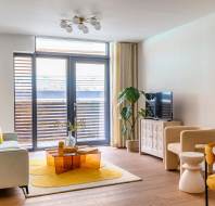 Modern living room with large windows, a light sofa, orange coffee table, yellow rug, and a TV on a white cabinet. A round dining table with yellow chairs is in the corner.