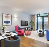 Modern living room and dining area with a blue sofa, pink chair, dining table set for four, and a wall-mounted TV. Large windows with curtains provide natural light. A plant decorates the corner.