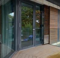 A glass door opens to a small balcony with wooden decking and a glass railing. Vertical wooden slats line part of the building's exterior. Trees are visible in the background.