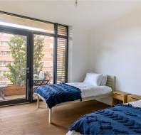 A bright bedroom with two single beds covered in blue quilts, a wooden desk, large window, and balcony view.