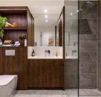 Modern bathroom with a wooden cabinet, white sink, and glass shower enclosure. Gray tiled walls and floor. Toilet paper is on a holder beside a white toilet. Shelves hold plants and toiletries.