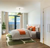 A modern bedroom with a white bed and orange accents, a green rug, wooden flooring, a hanging light, a potted plant, and a door leading to a balcony.