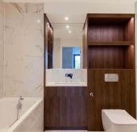 Modern bathroom with marble tiles, a bathtub, and wooden cabinetry. Reflective mirrors above the sink, next to a toilet. Recessed lighting illuminates the space.