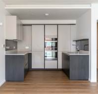 Modern kitchen with wood floor, dark cabinets, white countertops, built-in appliances, and stainless steel fixtures. Bright lighting and an open doorway are visible.