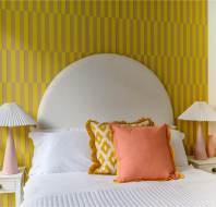 A bed with white linens, pink and yellow pillows, flanked by two white side tables with pink lamps. The wall features a yellow striped pattern with an arched white headboard.