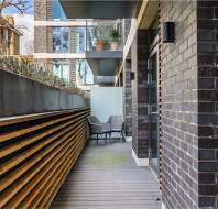 A modern balcony with two gray chairs and a small table. Wooden fencing on the left, brick wall on the right, and potted plants at the far end.