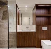 Modern bathroom with gray tiled shower, wooden vanity with sink, large mirror, and white toilet. Brown shelving unit beside the toilet and recessed lighting above.
