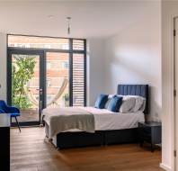 Modern bedroom with a large window, double bed, blue chair, and a hammock visible outside. White walls, wooden floor, and built-in closets complete the minimalist decor.