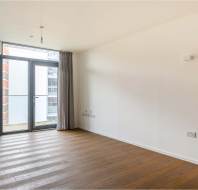 An empty room with light wooden flooring, white walls, and a floor-to-ceiling window with gray curtains leading to a balcony.