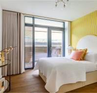 Modern bedroom with a large window, yellow accent wall, and a neatly made bed. A round mirror and floral arrangement are on a small glass table.