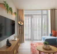 A modern living room with a blue sectional sofa, red and orange cushions, a mounted flat-screen TV, and a floor lamp. Green plants and framed art decorate the space. Natural light streams in.