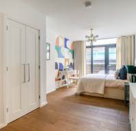Modern bedroom with wood flooring, a bed with light bedding, a desk with chair, and a colorful geometric wall. Large windows with curtains allow natural light. White cabinets are on the left.