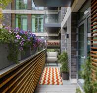 Modern narrow balcony with potted plants, a zigzag-patterned rug, and a cushioned chair against brick and glass building.
