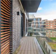 Balcony with wooden flooring and glass railing, flanked by a brick building with wooden slats on the left. Overlooks modern apartment buildings and a green rooftop under a blue sky.