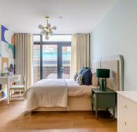 Modern bedroom with a bed, green nightstand, and geometric-patterned accent wall. Desk with a chair in the corner, light wood flooring, and large windows with beige curtains.