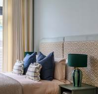 A modern bedroom with a large bed, several cushions, a green bedside lamp on a small table, and a window with curtains partially open.