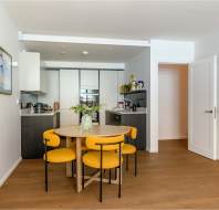 Modern kitchen with white walls, wooden floor, round table with yellow chairs, open shelving, and decorative wall art. Kitchen appliances and storage cabinets visible in the background.