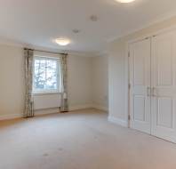 Empty room with beige carpet, white walls, and a window with floral curtains. Two closed white doors are on the right side. Ceiling lights are turned on.