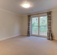 An empty room with beige carpet, white walls, a ceiling light, and large patio doors with curtains leading to a balcony.
