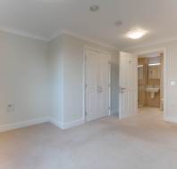 An empty room with beige carpet and walls, double doors open to a bathroom with a sink and mirror. Ceiling light and hallway visible.
