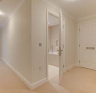 A hallway with beige carpet leads to an open bathroom on the right and a closed white door on the left. Natural light filters through a window at the end of the corridor.