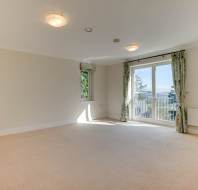 Empty living room with beige carpet, two ceiling lights, and green curtains. A window and glass doors lead to a balcony, and a radiator is under the right window. Wall and door are light-colored.