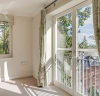 Bright room with light-colored walls and floral curtains. Sunlight streams through a window and French doors that open to a view of trees and a neighboring building.