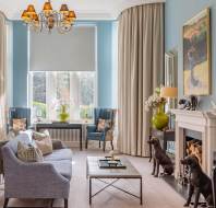 Elegant living room with blue walls, chandelier, three large windows, a sofa, two armchairs, a fireplace, and dog statues. A painting and vases decorate the space.
