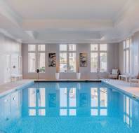 Indoor swimming pool with clear blue water, surrounded by beige tiles. Large windows let in natural light. Several lounge chairs and tables are placed along the sides.