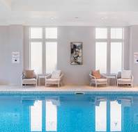 Indoor swimming pool with clear water, surrounded by white walls and large windows. Two chairs with cushions and a wall art piece are visible on the right side.