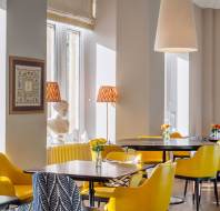 A well-lit dining area with yellow chairs, brown tables, and small flower arrangements. A statue is near the window, and art is displayed on the walls.