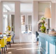 Bright cafe interior with yellow chairs, wooden tables, and a blue counter. Large windows allow natural light, while decorative vases and flowers add charm.