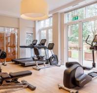 Modern gym with treadmills, exercise bikes, weight bench, and large windows letting in natural light.