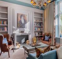 A cozy living room with bookshelves, a fireplace, and armchairs, featuring a large window and a chandelier.
