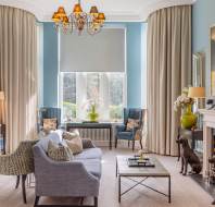 Elegant living room with blue walls, large windows, and beige curtains. Features a chandelier, fireplace, sofas, armchairs, and decorative sculptures. A framed painting hangs on the wall.