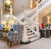 Elegant staircase in a well-lit room with two patterned armchairs and lamps on a console table, art on the wall, and a reception desk nearby.