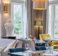 A modern dining room with chairs, tables set with glassware, and bright natural light from large windows. Vases and lamps decorate the space.
