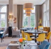Elegant dining room with tables set for service, featuring gray and yellow chairs, large pendant lights, art, and large windows with garden views.