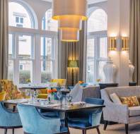 Elegant restaurant interior with arched windows, blue and yellow seating, and tables set with glassware. A contemporary chandelier hangs above, and a framed artwork is on the wall.