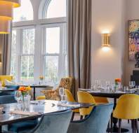 Elegant restaurant interior with colorful chairs, tables set with glassware and napkins, bright flowers, large windows, and art on the walls. Natural light fills the room.