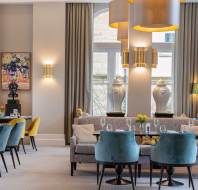 Elegant dining area with blue and yellow chairs, a gray sofa, and round tables set with glasses and flowers. Large windows, artwork, and vases decorate the room. Modern lighting overhead.