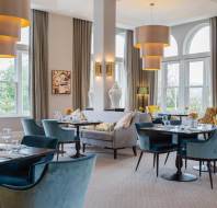 Elegant restaurant interior with blue chairs, round tables set with glassware, large pendant lights, and tall arched windows. A sofa and decorative items are visible in the background.