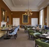 Elegantly decorated dining room with brown walls, round and square tables, assorted chairs, chandeliers, and framed artwork. Tables are set for dining.