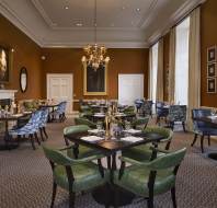 Elegant dining room with green and blue chairs, set tables, chandeliers, and paintings on warm brown walls. Natural light streams through large windows with beige curtains.