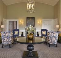 Elegant sitting room with a central round table, floral arrangement, patterned armchairs, green sofas, and decorative paintings on beige walls, lit by a large hanging lantern.