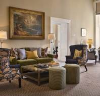 A cozy living room with a green sofa, patterned chairs, and two ottomans. A large painting hangs on the wall. Two lamps illuminate the space, which has large windows and a neutral color palette.