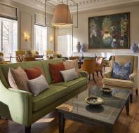 Elegant lounge with a green sofa, coffee table, and chairs. Large painting on the wall with decorative vases. Sunlight filters through tall windows.