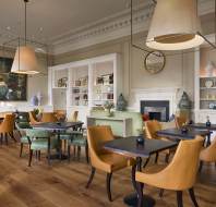 A modern cafe with brown and green chairs, wooden tables, large pendant lights, and wall art. Shelves display decorative vases.